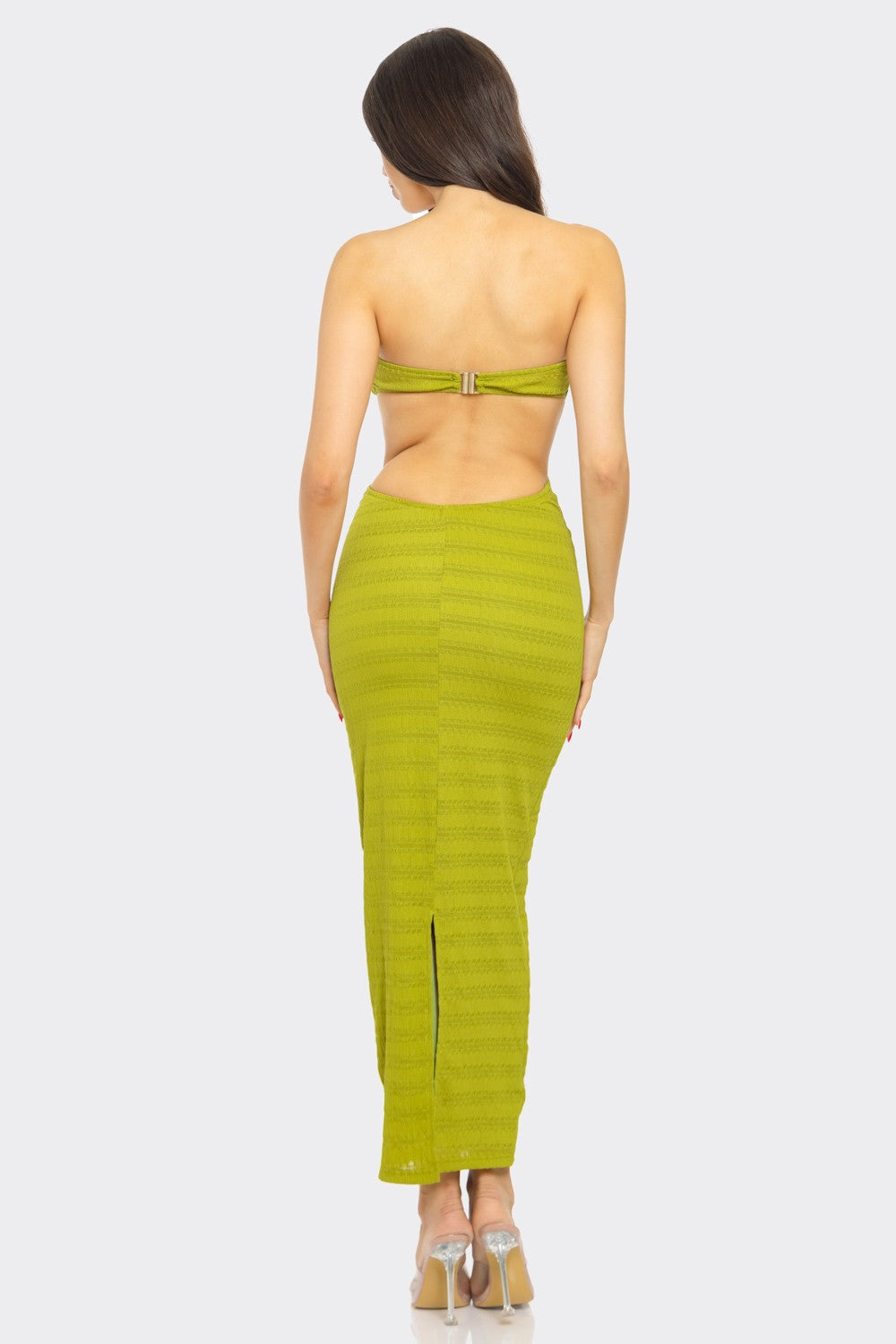 A woman with long dark hair stands with her back to the camera, wearing a bright yellow strapless maxi dress with an open back and a slit at the bottom. She pairs the dress with clear high heels. 