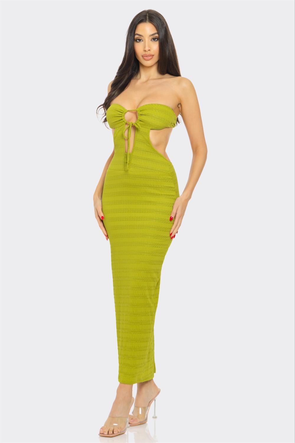 A woman with long dark hair poses against a white background, wearing a bright yellow strapless maxi dress with a front tie detail and cutout sides. She pairs the dress with clear high heels. The outfit accentuates her figure, showcasing a bold and seductive style.