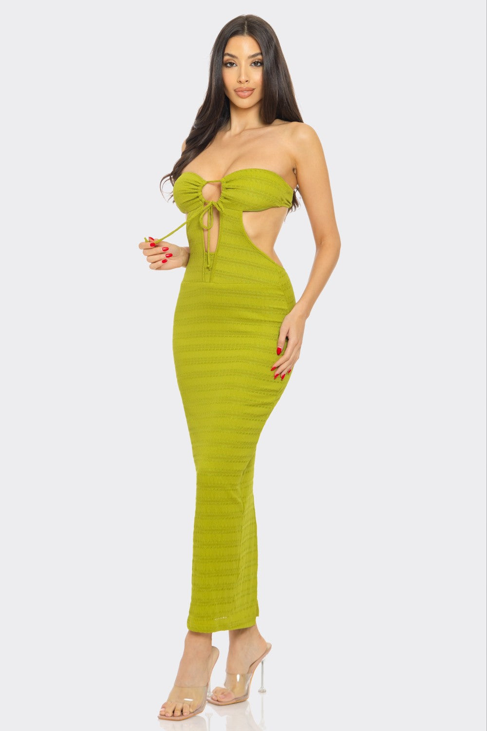 A woman with long dark hair poses against a white background, wearing a bright yellow strapless maxi dress with a front tie detail and cutout. She pairs the dress with clear high heels.
